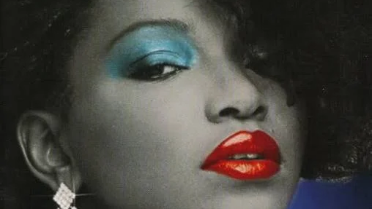 A black and white portrait of Shannon, styled with bright blue eyeshadow, vivid red lipstick, and a large rhinestone earring.